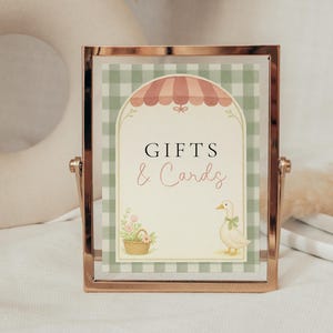 May include: A framed sign with the words "GIFTS & Cards" in black and pink script. The sign features a green and white checkered background, a pink awning, a basket of flowers, and a cartoon duck with a green bow. The frame is gold.