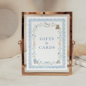 May include: A rectangular rose gold metal frame holding a white sign with the text GIFTS & CARDS in blue. The sign features a light blue scalloped border and illustrations of a rocking horse, teddy bear, rabbit, duck, baby carriage, and blocks.