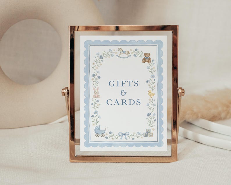 May include: A rectangular rose gold metal frame holding a white sign with the text GIFTS & CARDS in blue. The sign features a light blue scalloped border and illustrations of a rocking horse, teddy bear, rabbit, duck, baby carriage, and blocks.