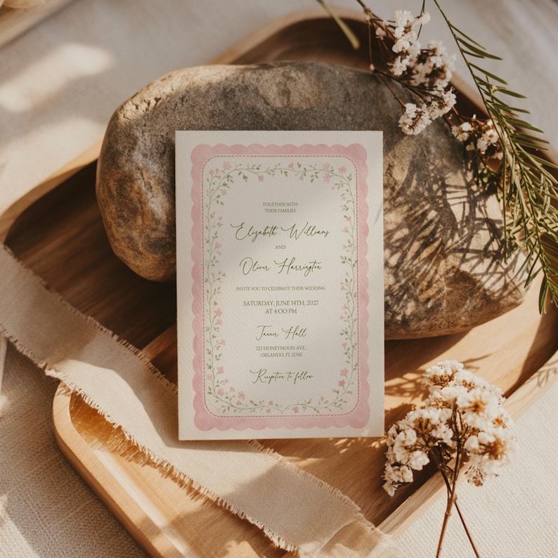 May include: A wedding invitation with a pink floral border and text. The invitation is on a wooden tray with a stone and floral accents. The text includes names, date, time, and location.