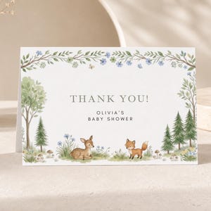 Peut inclure: Une carte de remerciement rectangulaire pour une fête prénatale avec une scène de forêt à l'aquarelle représentant un faon et un renard. Le texte indique THANK YOU! OLIVIA'S BABY SHOWER. La bordure inclut des arbres, des sapins et des fleurs.