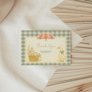 May include: A baby shower thank you card with a green gingham border. The card features a duck with a green bow, a basket of flowers, and an umbrella. The words "Thank You" are written in a script font.