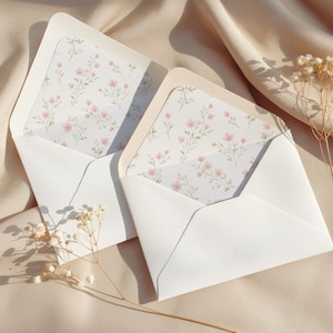Könnte beinhalten: Zwei weiße Umschläge mit floral gemusterten Innenfutter in Rosa und Grün sind geöffnet. Sie liegen auf beigem Stoff, mit Trockenblumen. Das Bild hat eine weiche, romantische Ästhetik, ideal für Schreibwaren.