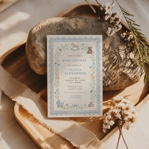 May include: A baby shower invitation with a blue floral border and illustrations of a rocking horse, teddy bear, bunny, duck, baby carriage, and blocks. The invitation reads "Join us for a Baby Shower in honor of Olivia Alexander, Saturday, August 24, 2028 at 3:00 PM."