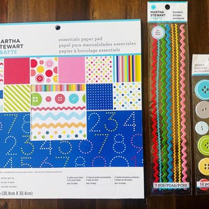 May include: A Martha Stewart Crafts essentials paper pad measuring 12 inches by 12 inches, a package of seven colorful zigzag borders, and a package of twelve assorted round buttons in various colors, all featuring printed branding and text.