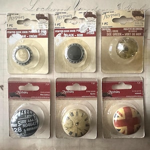 May include: Six individual 7gypsies brand knobs in retail packaging. The top row features a cream pewter knob, a black pewter knob, and a green crystal ball knob. The bottom row includes a black knob with white numbers, a clock face knob, and a Union Jack knob.