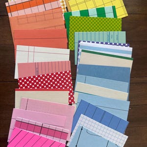 May include: A collection of colorful index cards and paper ephemera in various sizes and patterns. The cards feature stripes, grids, and geometric designs in shades of pink, blue, green, yellow, and orange. Some cards have punched holes.