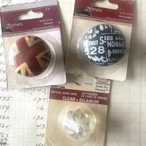 May include: Three decorative book knobs in their original packaging. One features a Union Jack design, another has black numbers on a white background, and the third is a clear crystal design.