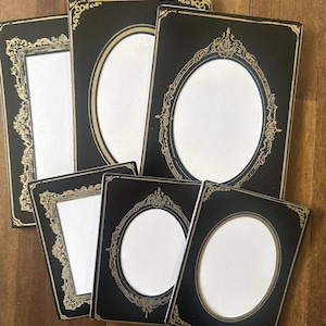 May include: A collection of black picture frames with gold ornate detailing. The frames have either rectangular or oval openings with white backgrounds. The frames are arranged on a wooden surface.