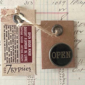 May include: A circular metal knob with a black center and the word OPEN in raised letters, attached to a rectangular brown paper tag. A second tag features the text OPEN BOOK KNOB, OUVERT FERMOIR DE LIVRE, and 7gypsies, connected by twine.