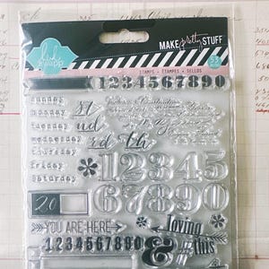 May include: A clear plastic package containing 53 clear photopolymer stamps by Heidi Swapp. The set includes numbers 0-9, days of the week, ordinal indicators, arrows, a small airplane, and phrases like "YOU ARE HERE", "loving", and "this".