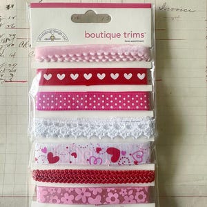 May include: A package of seven decorative ribbons by Doodlebug Design Inc. labeled boutique trims love assortment. The ribbons feature various patterns including pink velvet, red hearts, pink polka dots, white lace, hearts, red braid, and pink flowers.