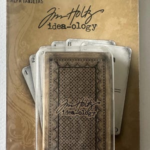 May include: A package of Tim Holtz Idea-ology Alpha Cards. The cards are in a clear plastic case. The cards have a vintage design with a brown and black color scheme. The packaging includes the text "ALPHA CARDS" and the brand name "Tim Holtz idea-ology".