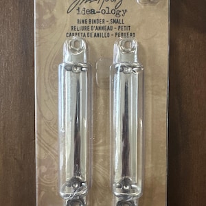 May include: Two small, shiny silver ring binders in a clear plastic package. The package has the text "Jim Holtz idea-ology RING BINDER - SMALL" and other text in French and Spanish. Also included are four brass fasteners.