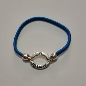 May include: A blue elastic bracelet featuring a silver-coloured circular charm. The charm is engraved with the word "BELIEVE". This bracelet is a simple, yet elegant accessory.