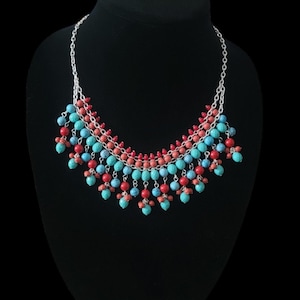 Op de afbeelding: Een zilveren ketting met turquoise, rode en koraalkleurige kralen. De ketting wordt getoond op een zwarte mannequin. De kralen zijn in een trapsgewijs patroon gerangschikt, waardoor een gelaagd effect ontstaat.