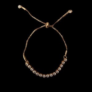 Könnte beinhalten: Goldfarbenes Armband mit einer Reihe kleiner, klarer Edelsteine. Das Armband hat eine zarte Kette und eine verschiebbare Perle zur Größenanpassung. Das Design ist schlicht und elegant, vor schwarzem Hintergrund.