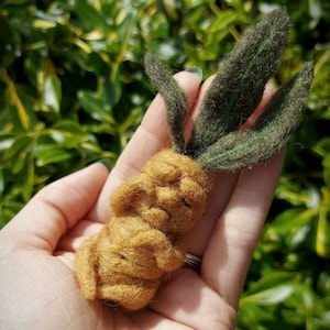 Puede incluir: Una escultura de fieltro marrón y verde hecha a mano que representa a una pequeña persona con una cabeza frondosa. La escultura está sostenida en la mano de una persona.