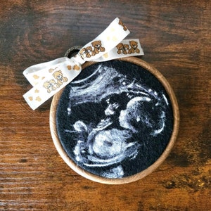 Puede incluir: Una imagen de ultrasonido en blanco y negro de un bebé en un marco de madera con una cinta blanca con ositos de peluche marrones impresos.