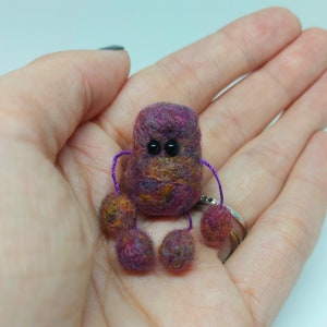 Puede incluir: Un pequeño robot de fieltro morado y naranja con ojos negros y brazos morados. El robot tiene cinco patas, cada una hecha de una bola de fieltro.