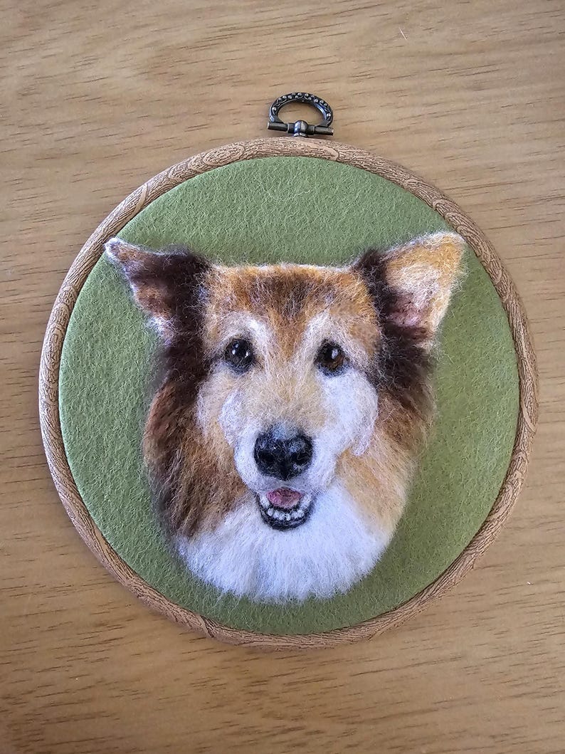 Puede incluir: Retrato de perro hecho a mano con fieltro de aguja en un bastidor de bordado redondo de madera. El perro tiene pelaje marr&oacute;n y blanco, una nariz negra y est&aacute; sobre un fondo de fieltro verde. El bastidor incluye un lazo met&aacute;lico para colgar.