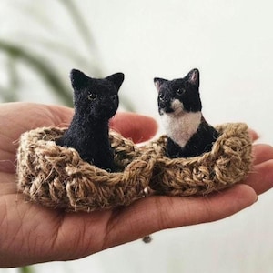 Puede incluir: Dos figuras de gatos en miniatura, una negra y otra negra y blanca, sentadas en cestas de yute tejidas separadas. Los gatos están detallados con rasgos realistas. Las cestas están conectadas, creando una pieza decorativa única.
