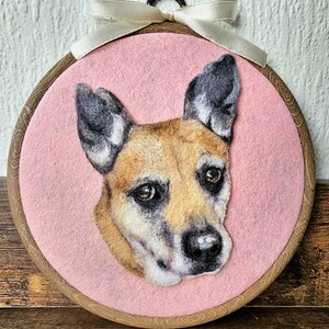 Puede incluir: Retrato de perro de fieltro en un aro de bordado de madera. El perro tiene pelaje marr&oacute;n y negro, sobre un fondo rosa. Una cinta color crema est&aacute; atada en la parte superior. La cara del perro es el foco, con detalles.