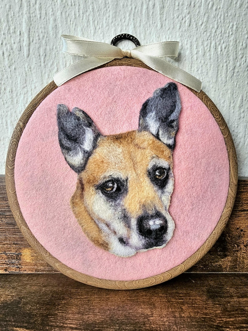 Puede incluir: Retrato de perro de fieltro en un aro de bordado de madera. El perro tiene pelaje marr&oacute;n y negro, sobre un fondo rosa. Una cinta color crema est&aacute; atada en la parte superior. La cara del perro es el foco, con detalles.