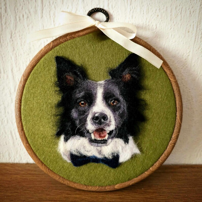 Puede incluir: Retrato de fieltro de un perro Border Collie, con pelaje blanco y negro y una pajarita azul. La obra de arte est&aacute; en un bastidor de bordado de madera, con un fondo verde y una cinta crema para colgar.