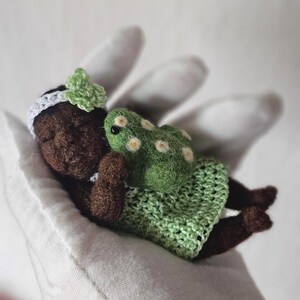 Puede incluir: Una muñeca en miniatura hecha a mano con piel morena, que lleva una diadema blanca con un lazo verde y un vestido de ganchillo verde. Una pequeña tortuga verde con manchas blancas descansa junto a la muñeca. La muñeca está acostada.