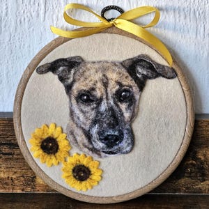 Puede incluir: Una obra de arte en fieltro enmarcada, que presenta un retrato de perro con pelaje atigrado, dos girasoles amarillos y una cinta amarilla. La obra est&aacute; sobre un fondo beige, dentro de un c&iacute;rculo de madera. Aproximadamente 20 cm de di&aacute;metro.