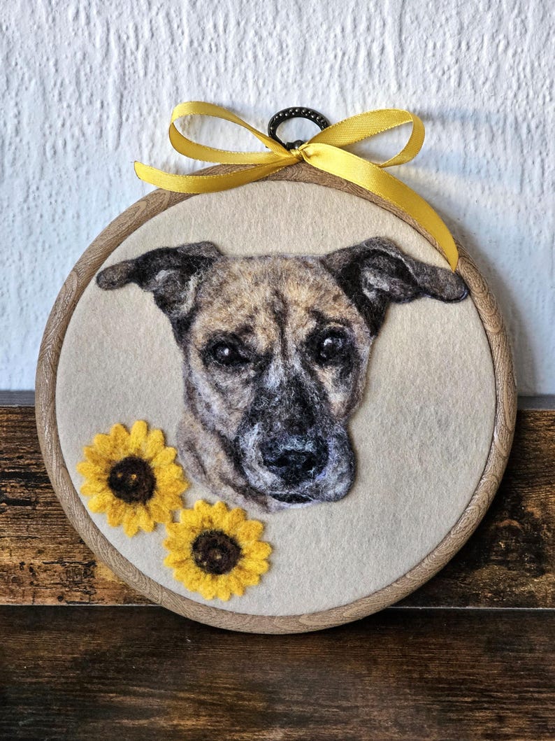 Puede incluir: Una obra de arte en fieltro enmarcada, que presenta un retrato de perro con pelaje atigrado, dos girasoles amarillos y una cinta amarilla. La obra est&aacute; sobre un fondo beige, dentro de un c&iacute;rculo de madera. Aproximadamente 20 cm de di&aacute;metro.