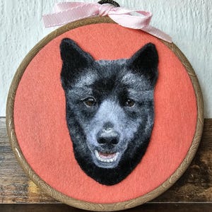 Puede incluir: Retrato afieltrado de un perro negro con hocico gris claro y ojos marrones, sobre un fondo coral en un bastidor de bordado de madera. Un lazo de cinta rosa est&aacute; atado en la parte superior.