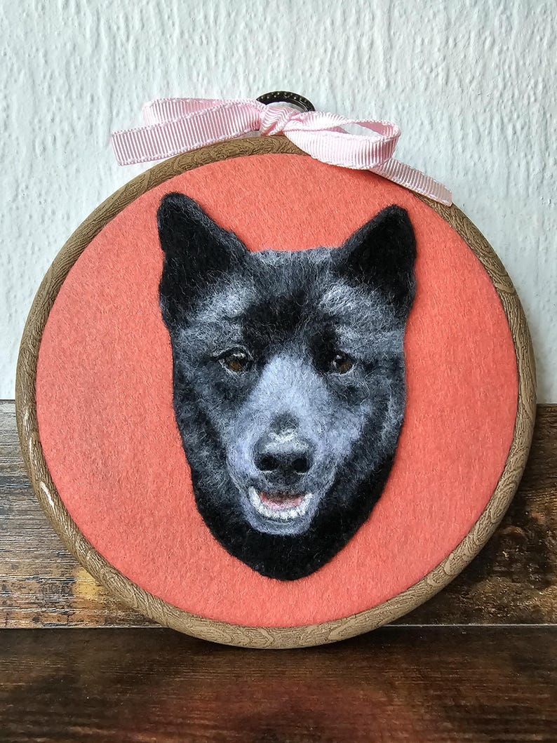 Puede incluir: Retrato afieltrado de un perro negro con hocico gris claro y ojos marrones, sobre un fondo coral en un bastidor de bordado de madera. Un lazo de cinta rosa est&aacute; atado en la parte superior.
