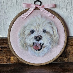 Puede incluir: Una pieza de arte textil redonda enmarcada, que presenta un perro claro y esponjoso con la lengua rosa. El retrato del perro est&aacute; sobre un fondo rosa, enmarcado por un aro de madera y una cinta rosa.