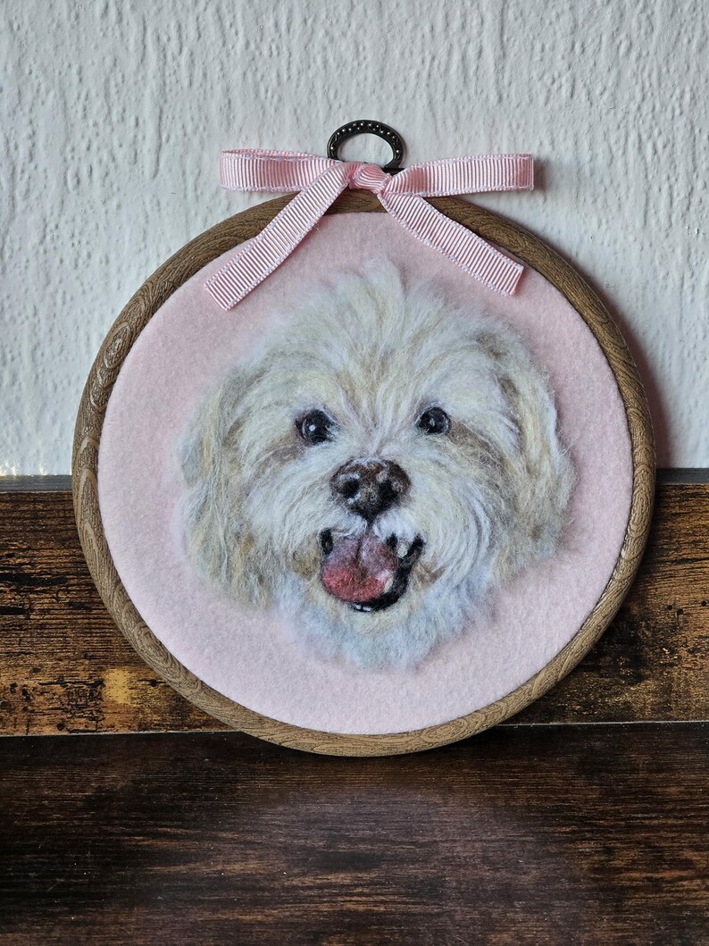 Puede incluir: Una pieza de arte textil redonda enmarcada, que presenta un perro claro y esponjoso con la lengua rosa. El retrato del perro est&aacute; sobre un fondo rosa, enmarcado por un aro de madera y una cinta rosa.
