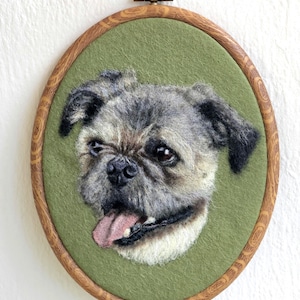 Puede incluir: Retrato textil enmarcado de forma ovalada, que representa un perro de fieltro con pelaje gris y marr&oacute;n, con la lengua fuera. El perro est&aacute; sobre un fondo verde, dentro de un marco de madera con un lazo met&aacute;lico para colgar.