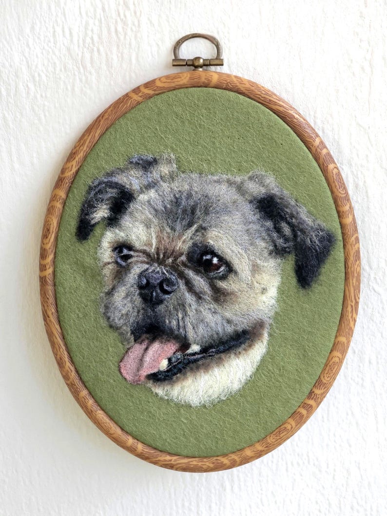 Puede incluir: Retrato textil enmarcado de forma ovalada, que representa un perro de fieltro con pelaje gris y marr&oacute;n, con la lengua fuera. El perro est&aacute; sobre un fondo verde, dentro de un marco de madera con un lazo met&aacute;lico para colgar.
