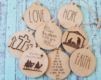 Op de afbeelding: Set van acht houten kerstversieringen met verschillende religieuze thema's. De versieringen zijn voorzien van gegraveerde ontwerpen en tekst, waaronder "LOVE", "HOPE", "FAITH", "Wise Men Seek Him Still", "For I know the plans I have for you - Jeremiah 29:11", en een afbeelding van de kerststal.