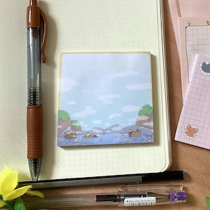 Może przedstawiać: Zestaw czterech różowych i białych, kratkowanych bloczków samoprzylepnych z uroczymi ilustracjami kotów. Bloczki leżą na drewnianej powierzchni z notesem i długopisem.