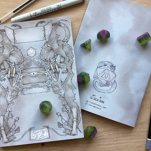 Può includere: Due copertine di quaderno vuote con un'illustrazione di un drago e un forziere del tesoro su una e un drago che tiene un dado a venti facce sull'altra. I quaderni sono su una superficie di legno con dadi verdi e viola sparsi intorno.