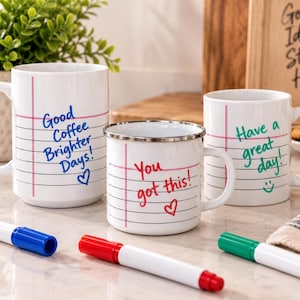 May include: Three white ceramic and metal mugs featuring a notebook paper design with horizontal lines and a vertical red margin. The mugs display handwritten-style text: Good Coffee Brighter Days!, You got this!, and Have a great day!.