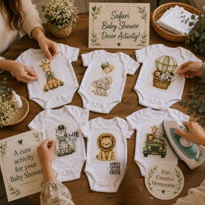 May include: Six white baby bodysuits featuring various safari animal illustrations including a giraffe, elephant, lion, and zebra. A sign reads Safari Baby Shower Decor Activity, and an iron is positioned next to the bodysuits on a wooden table.