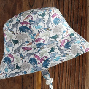 May include: A white bucket hat with a colorful print of animals, including elephants, giraffes, and birds. The hat has a chin strap and a wooden handle.