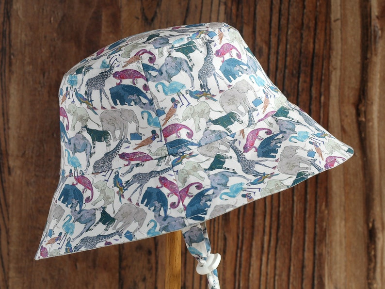 May include: A white bucket hat with a colorful print of animals, including elephants, giraffes, and birds. The hat has a chin strap and a wooden handle.
