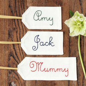 May include: Three white fabric gift tags with ribbon ties. The tags are embroidered with the names "Amy", "Jack", and "Mummy".