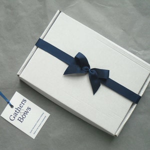 May include: A white gift box with a navy blue ribbon tied in a bow. The box has a label that reads "Gathers & Bows" and "Quality Handmade Children's Clothing Made in the Sussex Countryside".