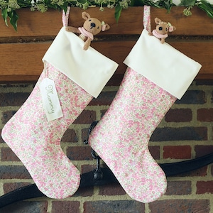 May include: Two pink and white Christmas stockings with floral patterns hanging on a brick fireplace mantel. The stockings have white felt cuffs and are decorated with small crocheted reindeer ornaments. One stocking has a tag that says "Mommy".