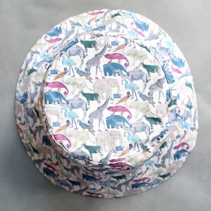May include: A white bucket hat with a colorful print of animals, including giraffes, elephants, and birds. The hat has a wide brim and a flat crown.