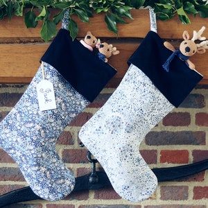May include: Two festive Christmas stockings hang from a wooden mantelpiece, adorned with greenery. One is patterned with blue florals, the other with white snowflakes. Each has a black cuff and a small crocheted reindeer.